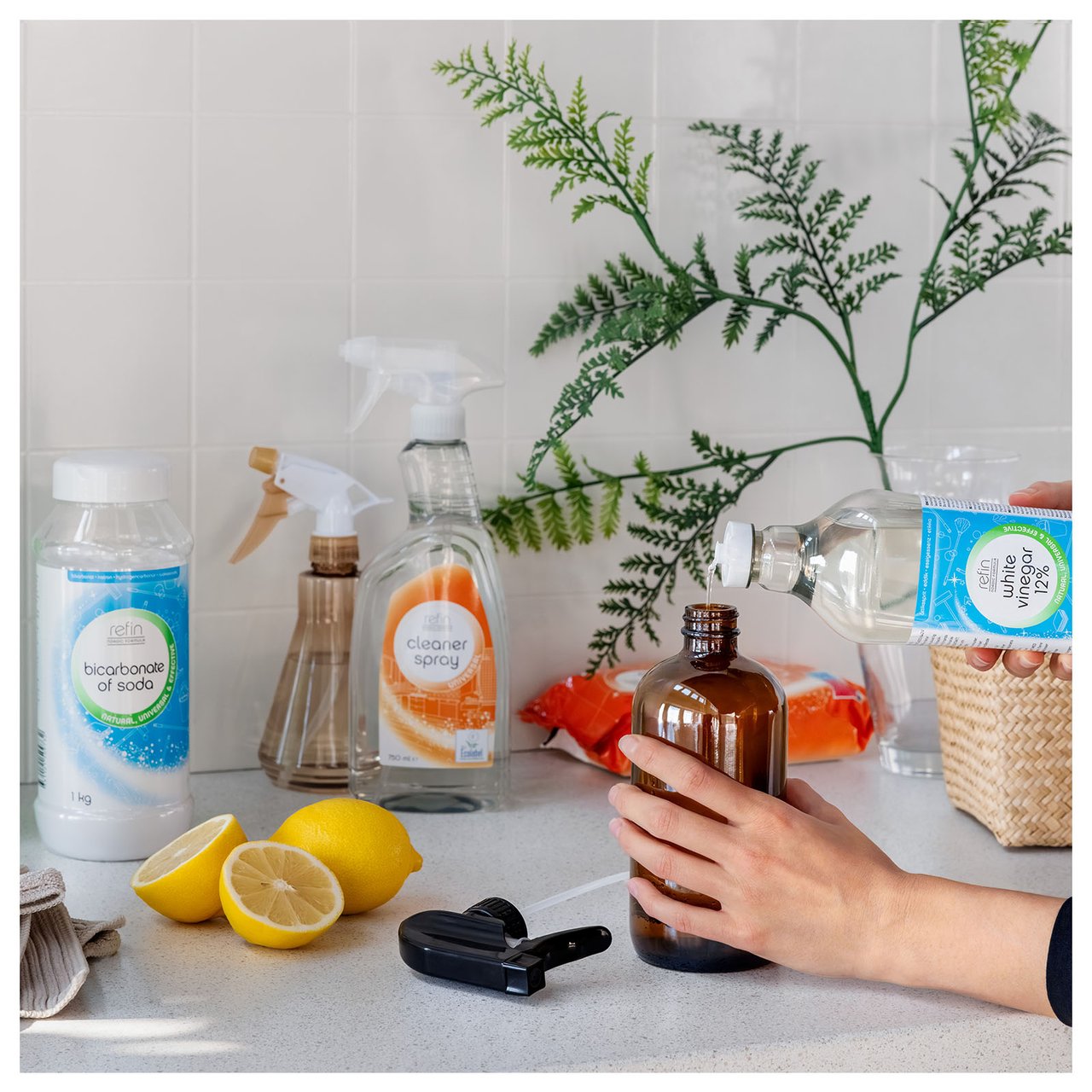 Hand, die Essig in eine braune Sprühflasche füllt. Im Hintergrund Natron, Reinigungsspray und Zitronen.
