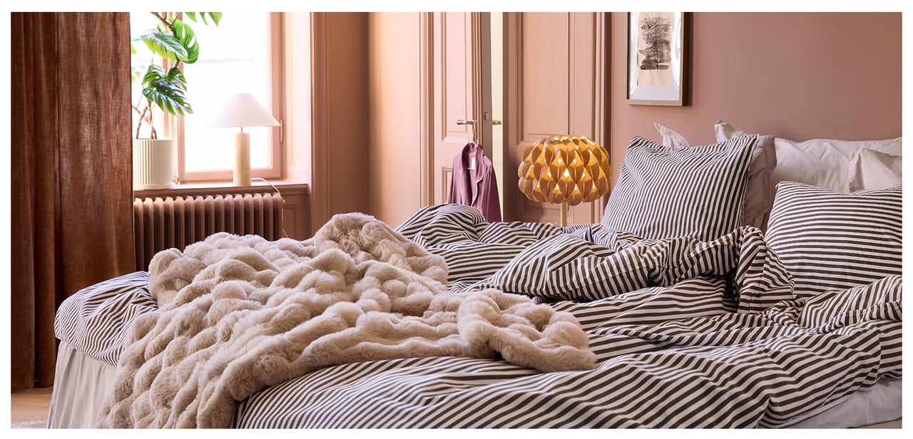 Ett sovrum med terrakottafärgade väggar, randiga sängkläder och en fluffig, beige pläd.