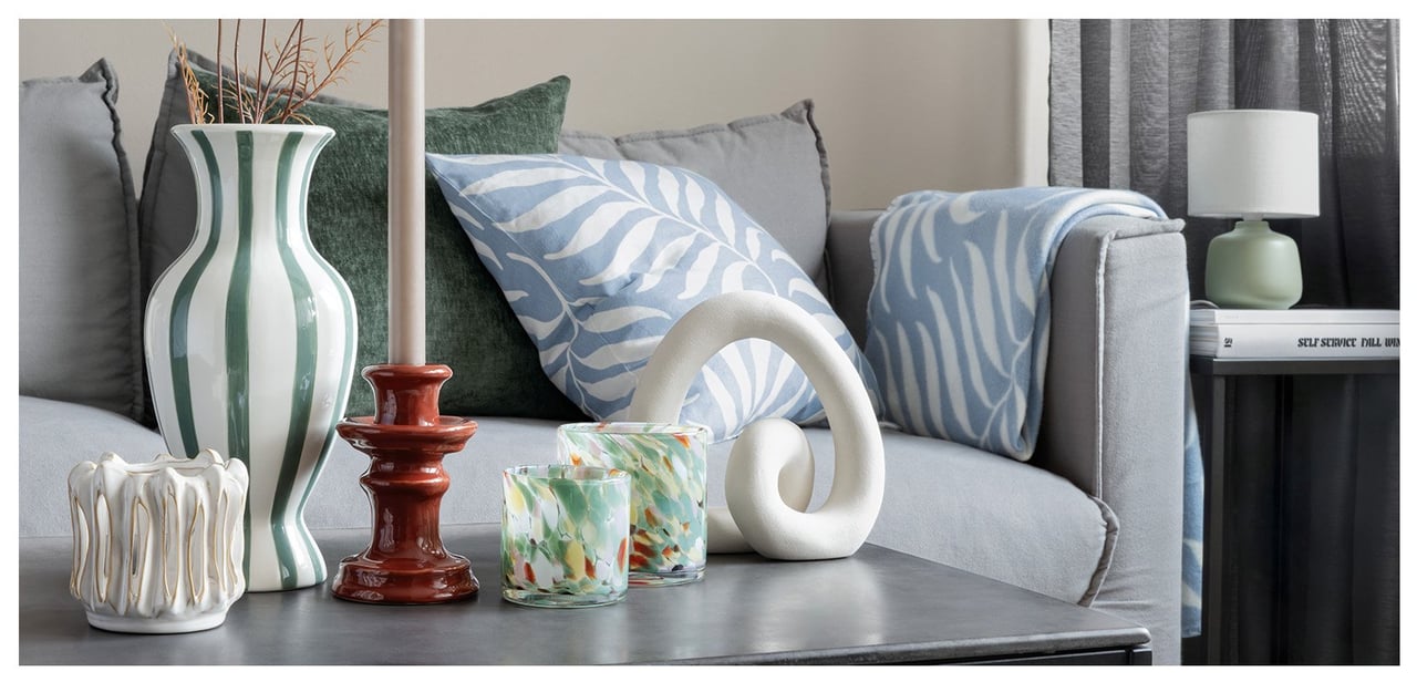 Teil eines Sofas und eines Couchtisches in einer gemütlichen Umgebung mit Kissen, Decken und Kerzenständern in Graugrüntönen.