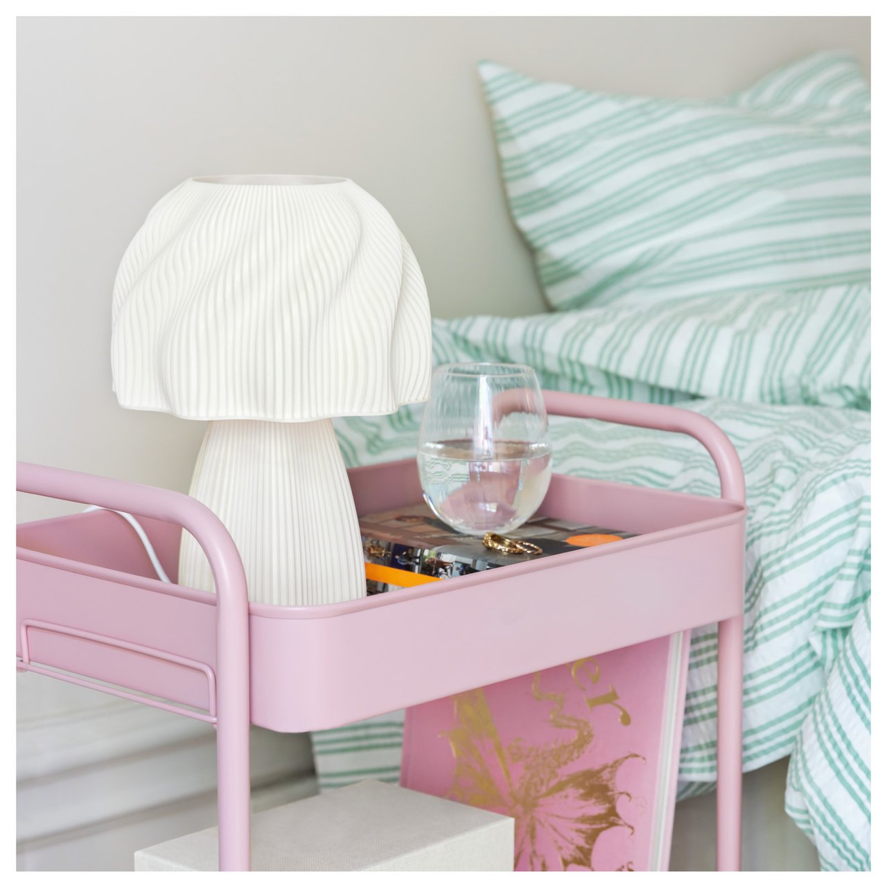 Kleine Tischlampe und Glas Wasser auf einem Rolltisch neben einem gemachten Bett.