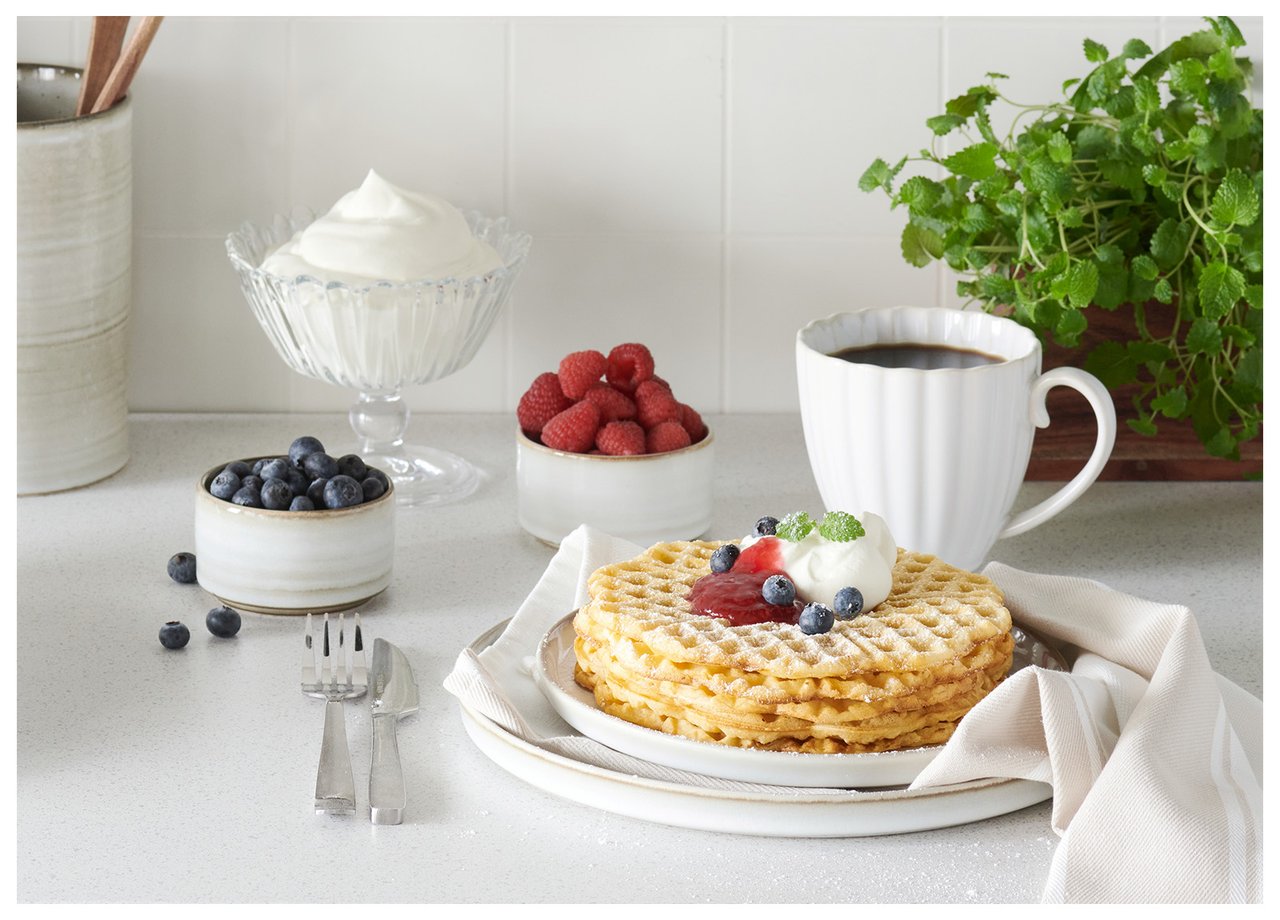 En hög med våfflor på en tallrik. Sylt, bär, vispgrädde och en kopp kaffe bredvid.