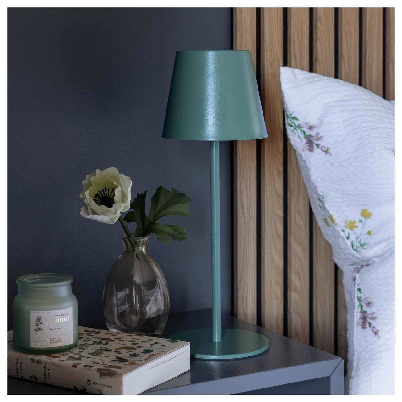 Bett mit geblümtem Kissenbezug neben einem Nachttisch mit schmaler Tischlampe, Duftkerze, Vase und Buch.