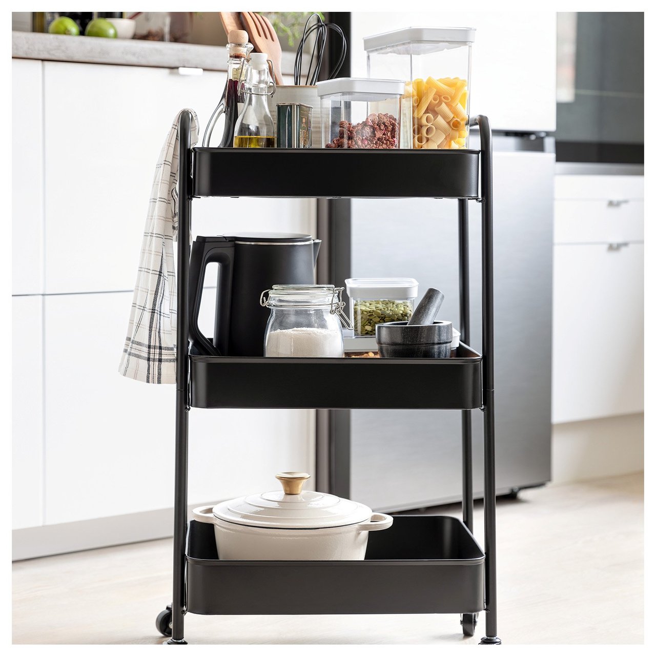 Schwarzer Rollwagen in weißer Küche. Auf dem Rollwagen stehen ein Topf, ein Mörser, ein Wasserkocher und Vorratsbehälter aus Kunststoff.