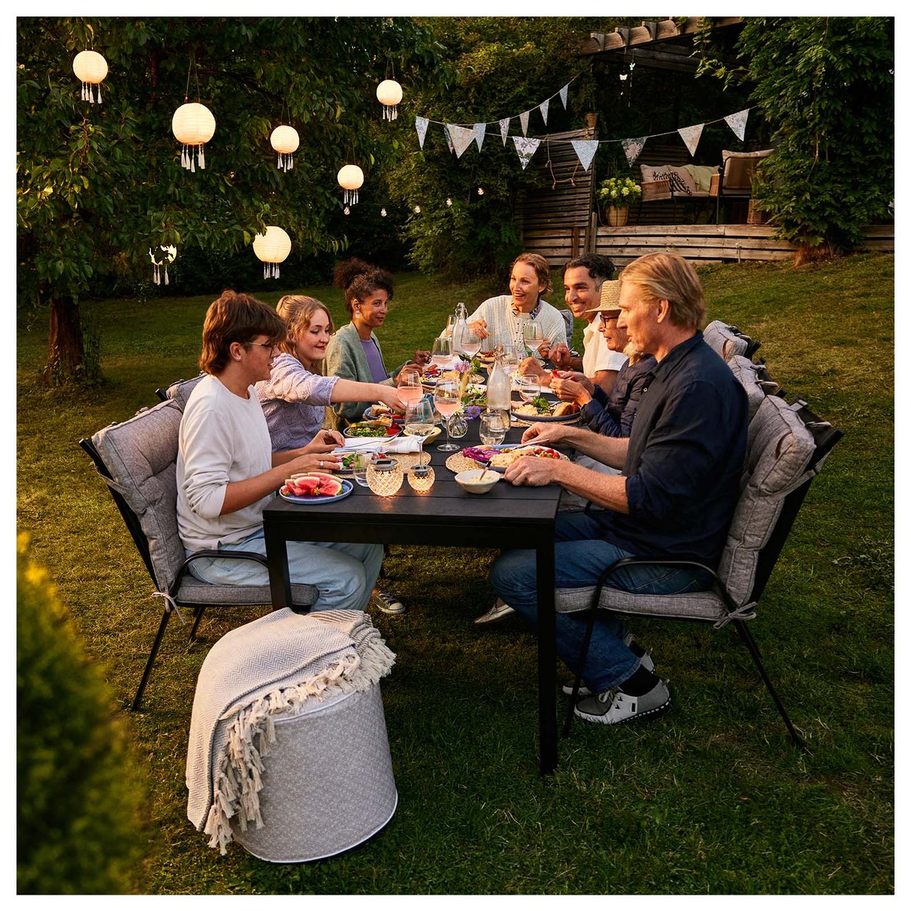 Sieben Personen essen im Freien in einem Garten zu Abend, der mit in der Dunkelheit leuchtenden Solarlichterketten geschmückt ist.