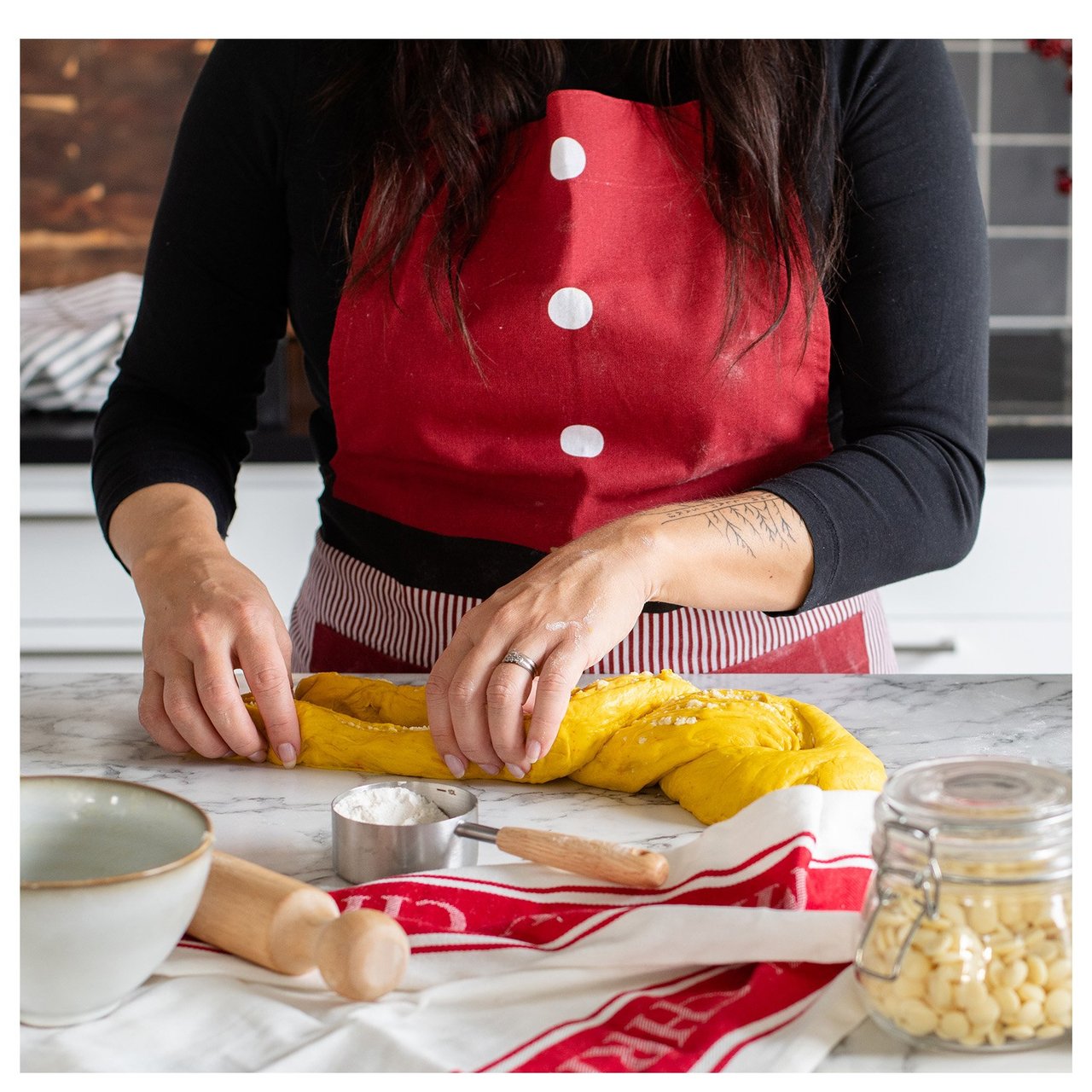 Eine Person in einer roten Schürze flicht Safranteig auf einer Küchenarbeitsfläche mit Backutensilien und Zutaten.
