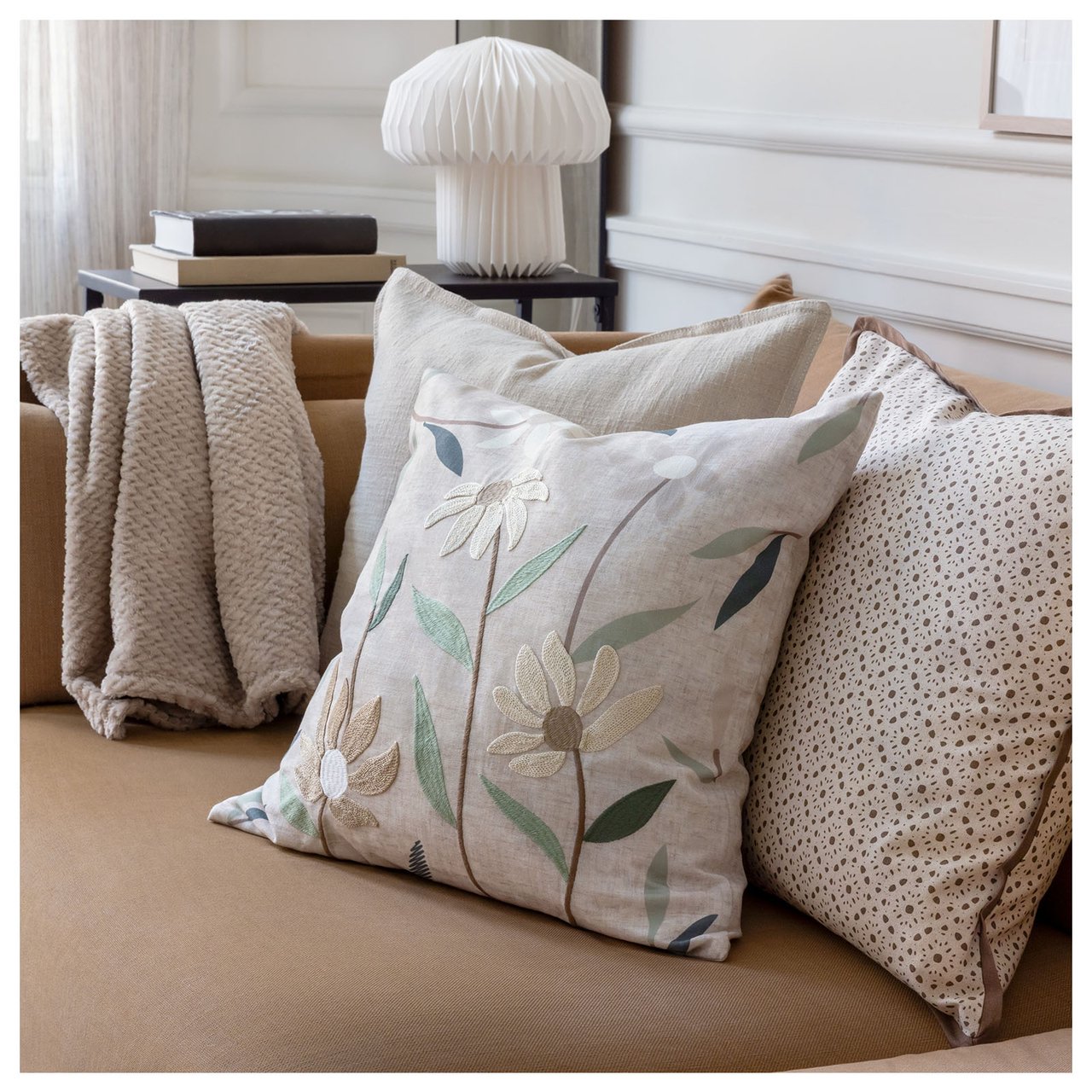 Tre naturfargede puter med forskjellige mønstre ligger i en sofakrok. Bak står det en hvit lampe på et sidebord.