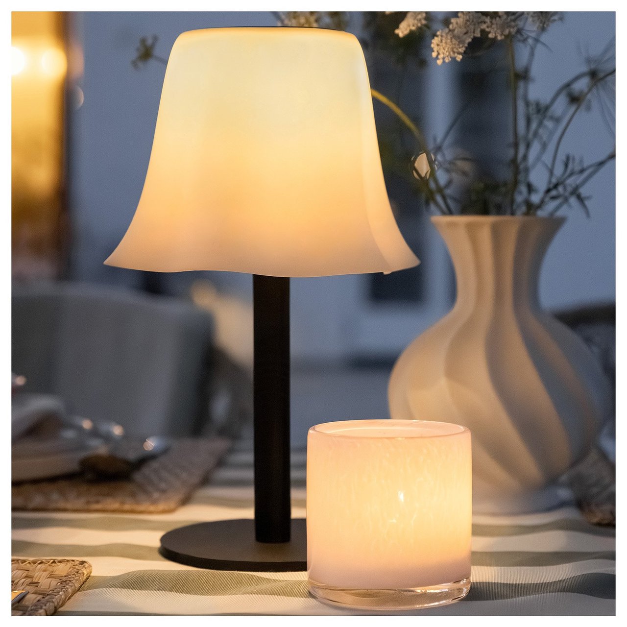 Solcellelampen Hemsön med svart fot og hvit, bølget skjerm på et bord sammen med en hvit telysholder.