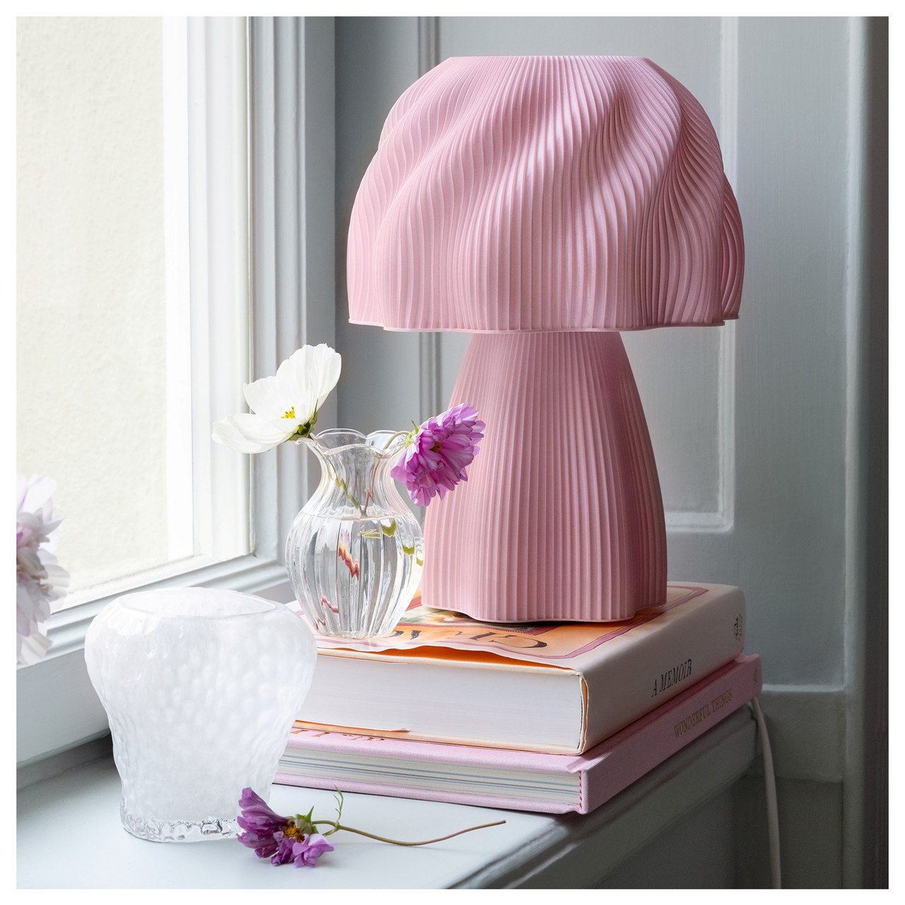 En rosa, bølgeformet bordlampe står på en stabel med bøker i et vindu. Ved siden av står det to mindre glassvaser.