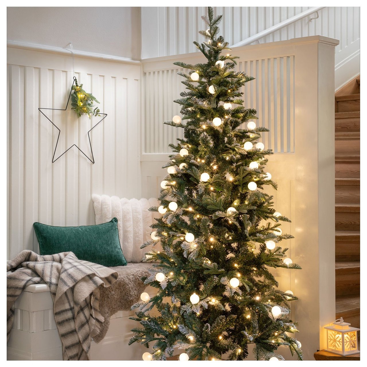 Mysigt hörn med julgran klädd i ljusslinga med glober, sittbänk med filt och kuddar, lykta på golvet och stjärna på väggen.