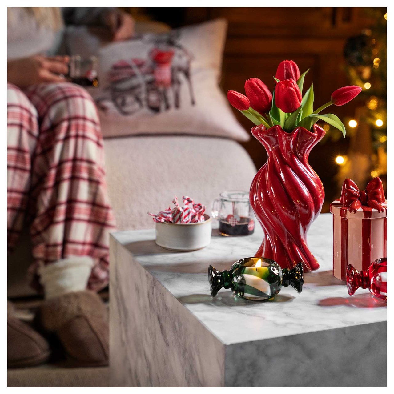 En person i pyjamas och tofflor sitter i en soffa. På bordet står röda tulpaner och karamellformade ljusstakar.