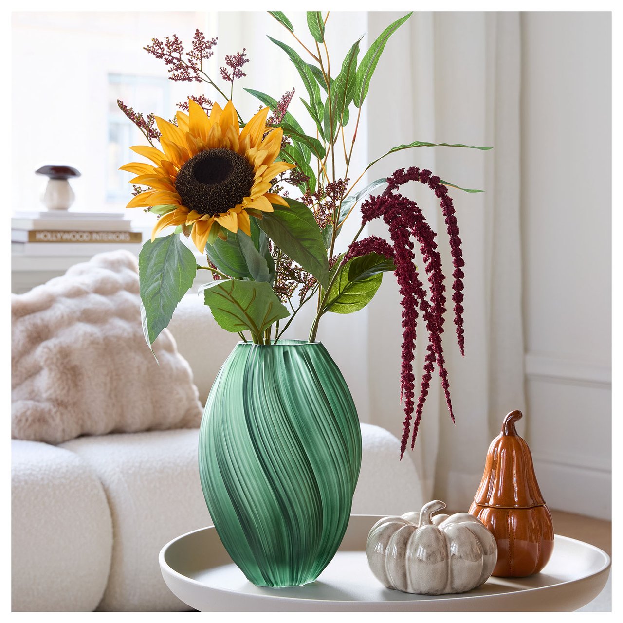 Grønn vase med solsikke, grønne kvister og rød amarant står på et lite beige bord. En sofa vises i bakgrunnen.