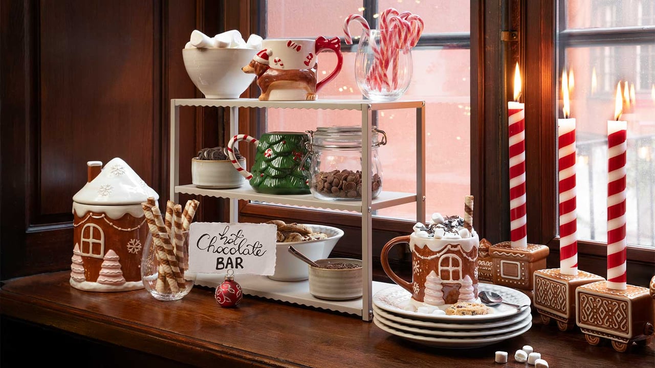 En hot chocolate bar uppdukad i ett fönster med julmuggar, marshmallows, polkagrisar och tända ljus.