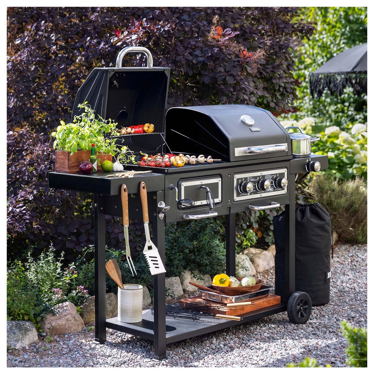 Kol- och gasolgrill med tillagade grillspett står på grusgång i trädgård.