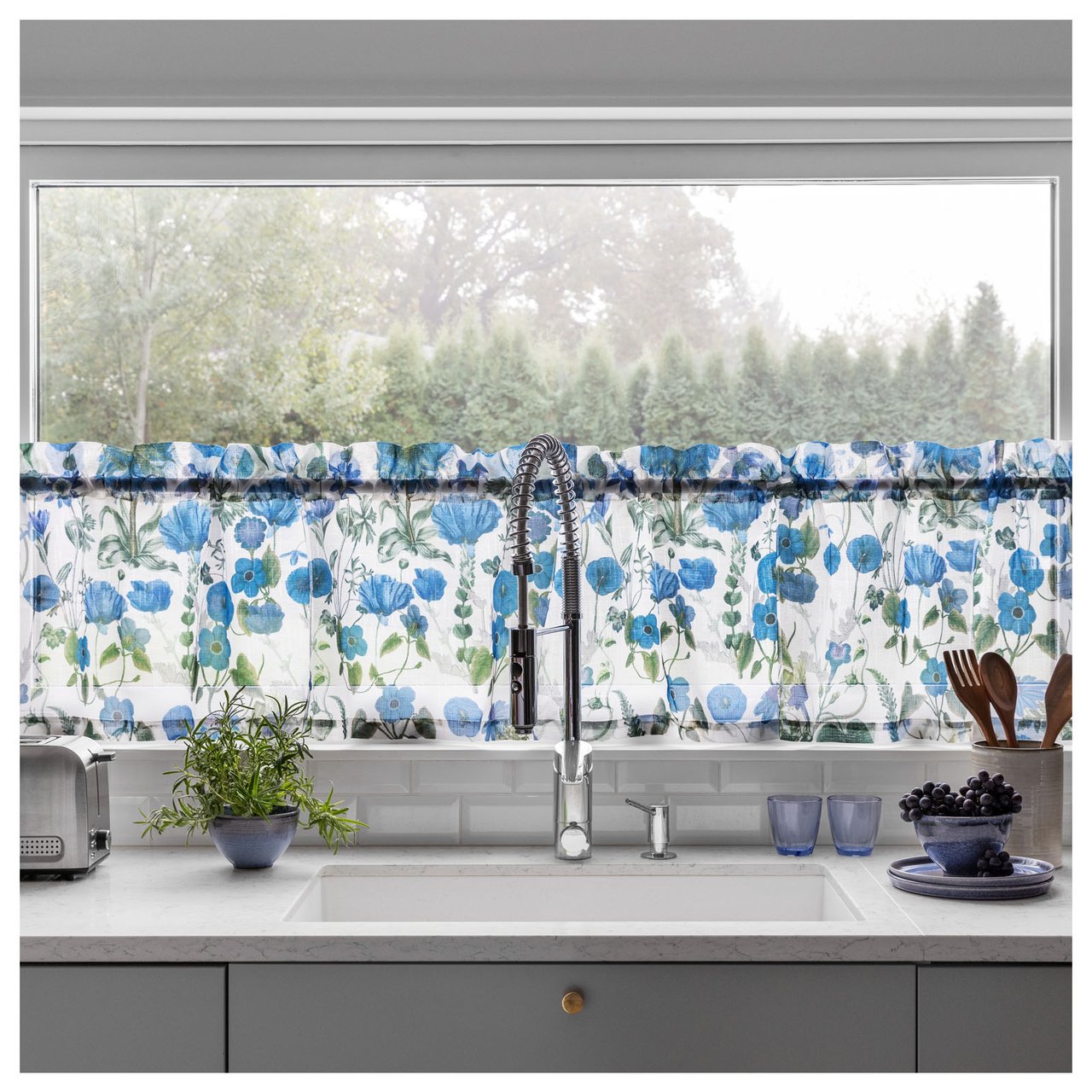 Keittiön ikkuna, jossa on sinikukkainen verhokappa tiskialtaan yläpuolella ja työtaso keittiövälineineen.
