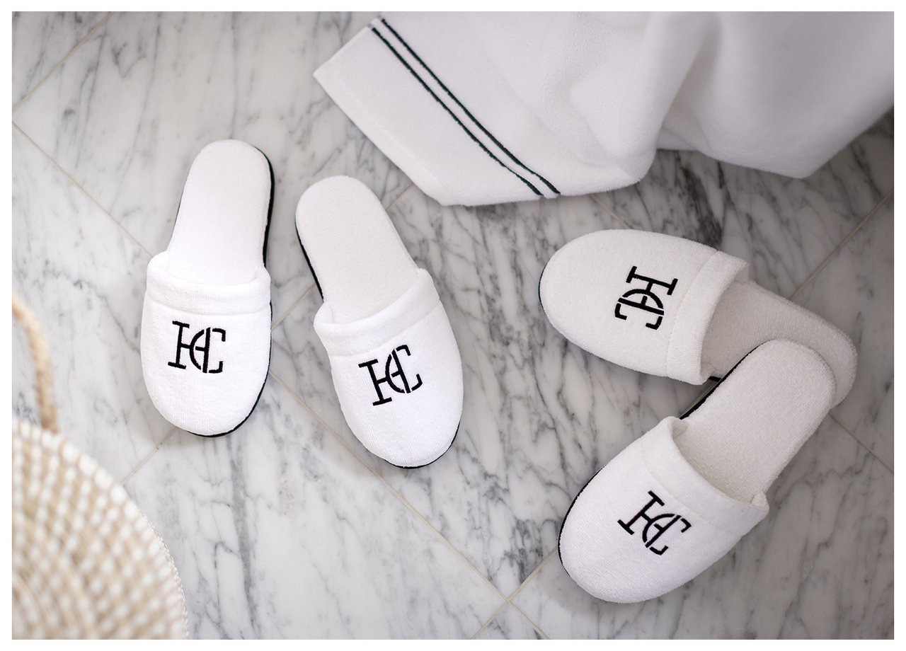 Zwei Paar weiße Hausschuhe mit schwarzem Emblem auf Marmorboden.