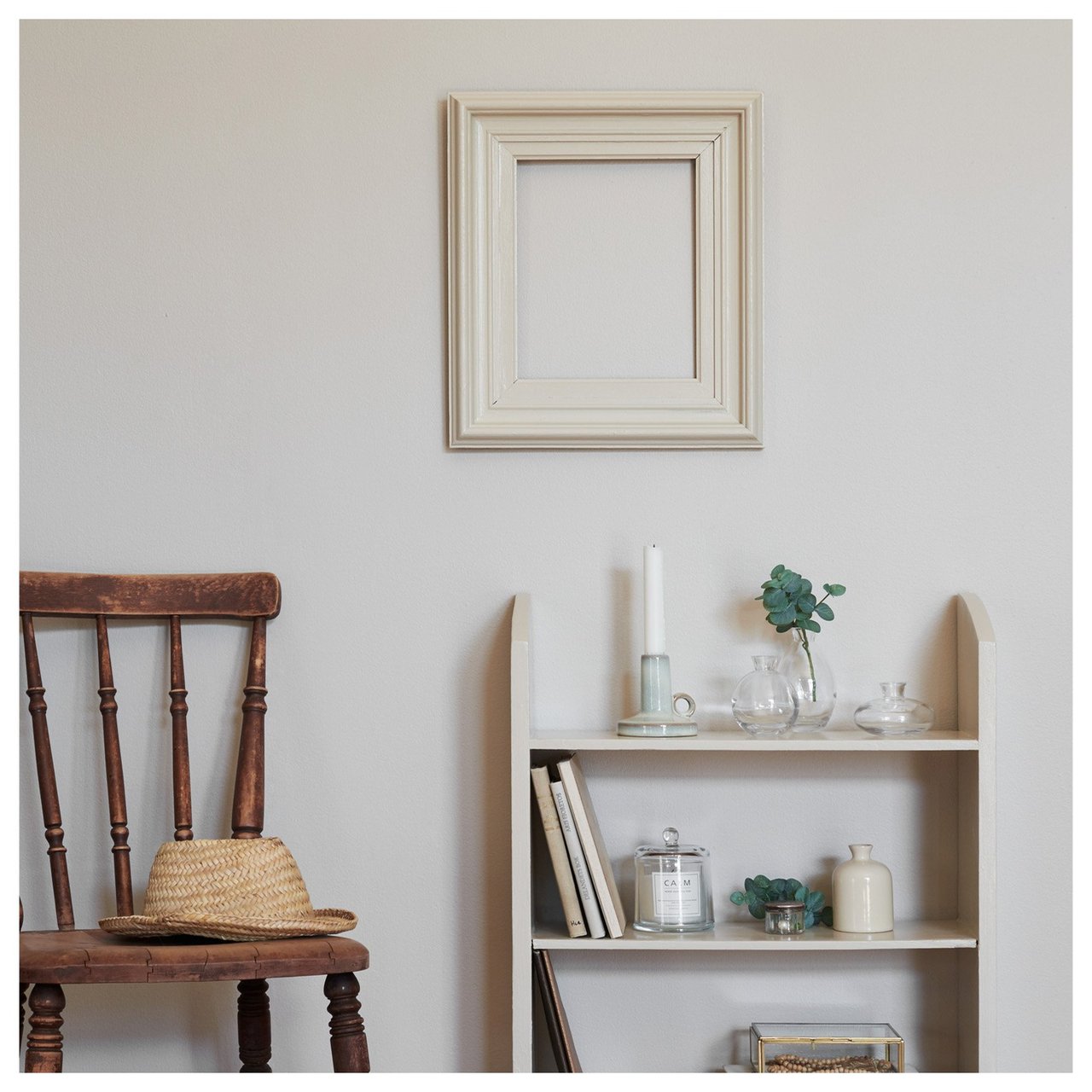 Innredning i naturhvite toner med hylle, rustikk trestol som det ligger en hatt på, samt en tom ramme på veggen.