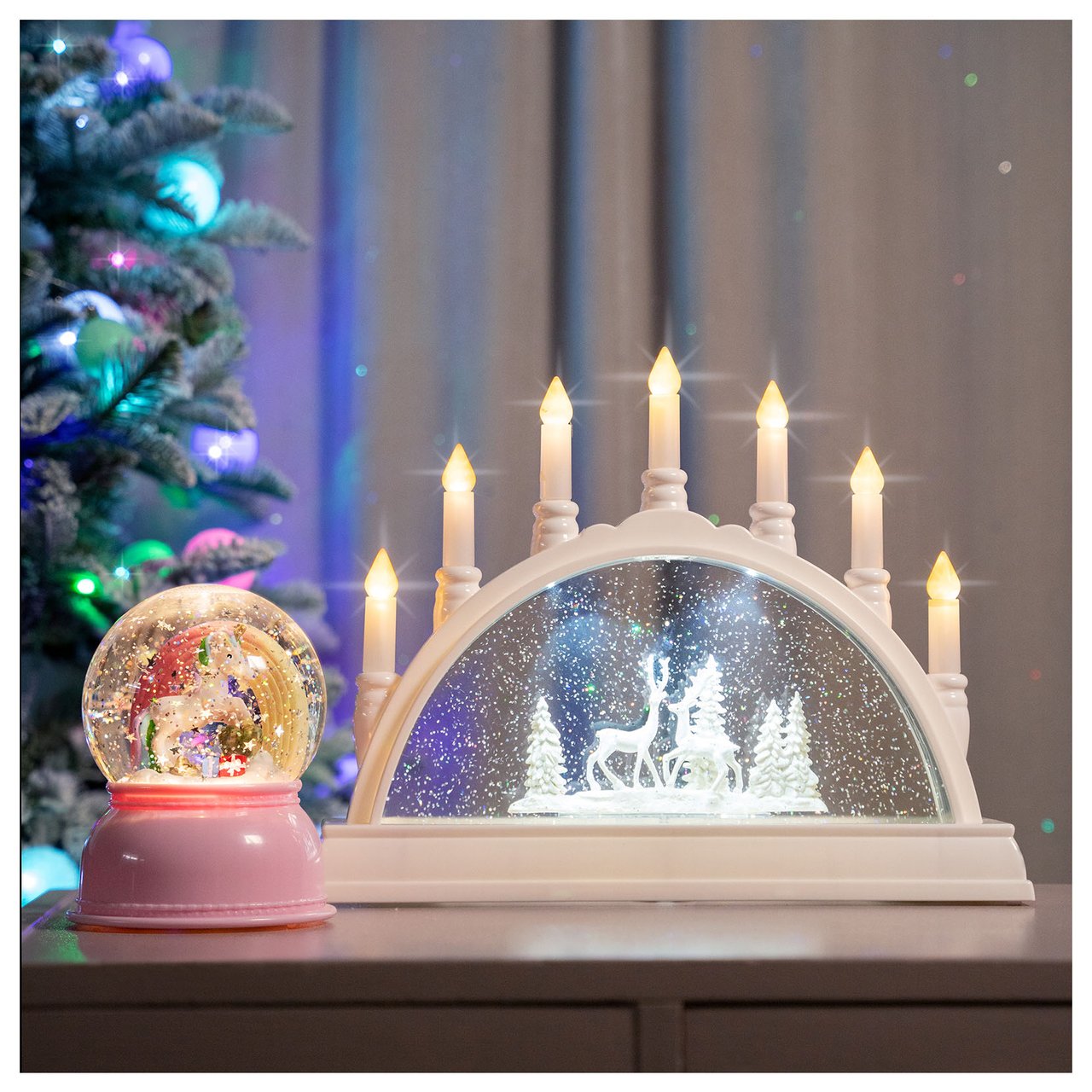 Tisch mit Adventsleuchter und Schneekugel vor einem beleuchteten Weihnachtsbaum.