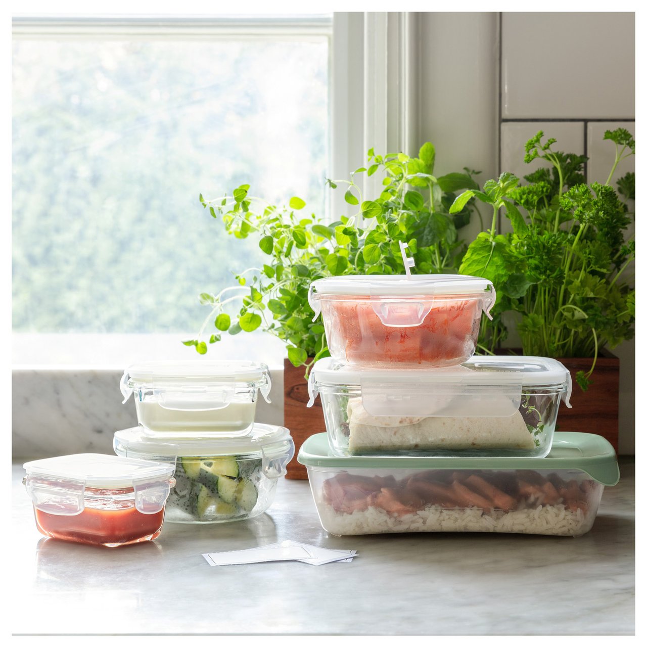 Matlådor med olika innehåll uppställda på köksbänk framför fönster med färska örter.