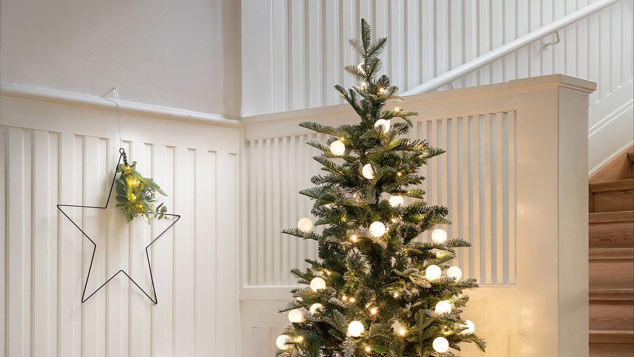 Ein schmaler Kunstbaum mit Beleuchtung und weißen Kugeln in einer festlich dekorierten Ecke bei einer Treppe.