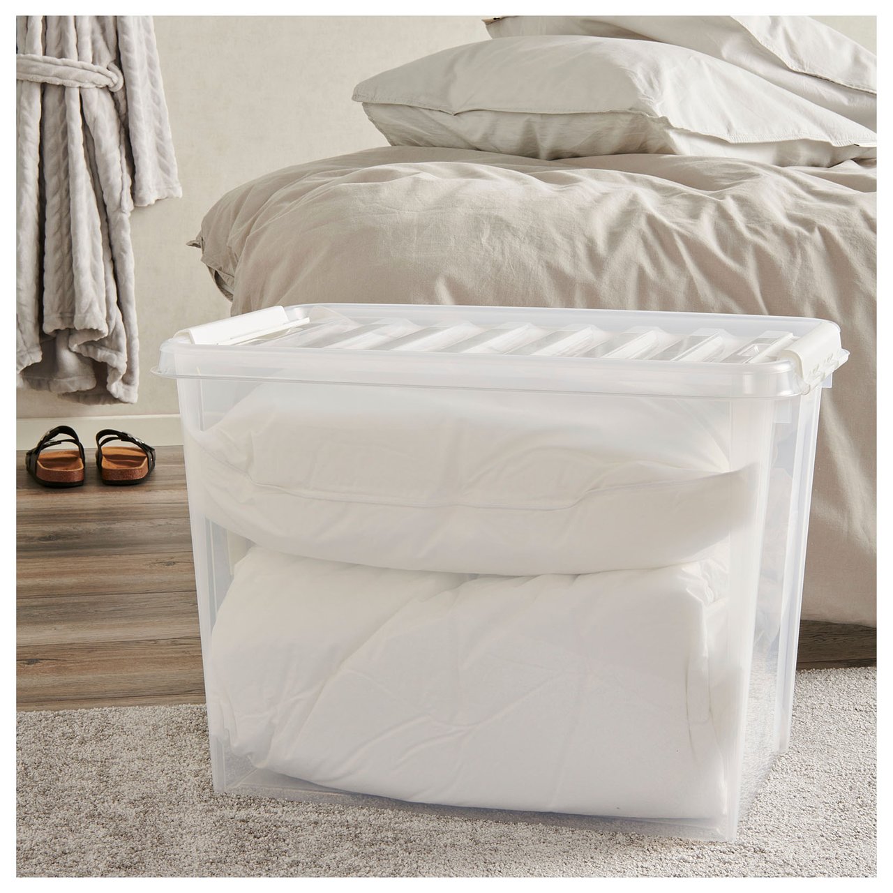 Große Kunststoffbox mit Kopfkissen, die auf einem Teppich vor einem Bett steht.