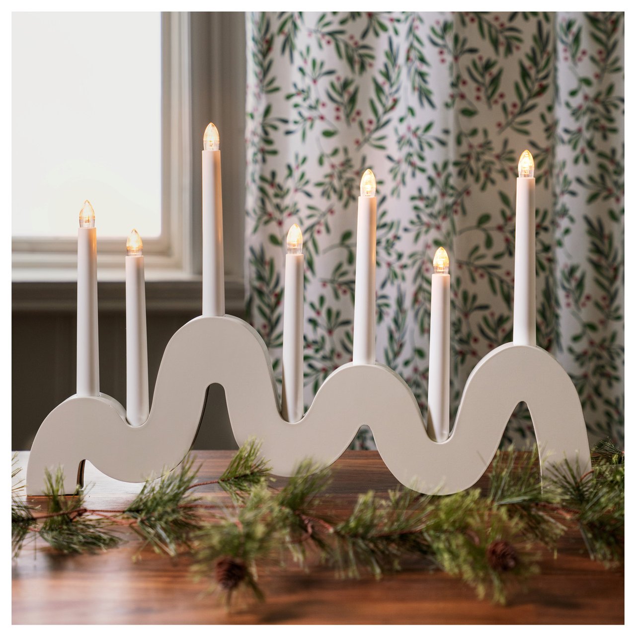 Ein weißer Adventsleuchter in geschwungenen Formen steht vor einem Fenster.