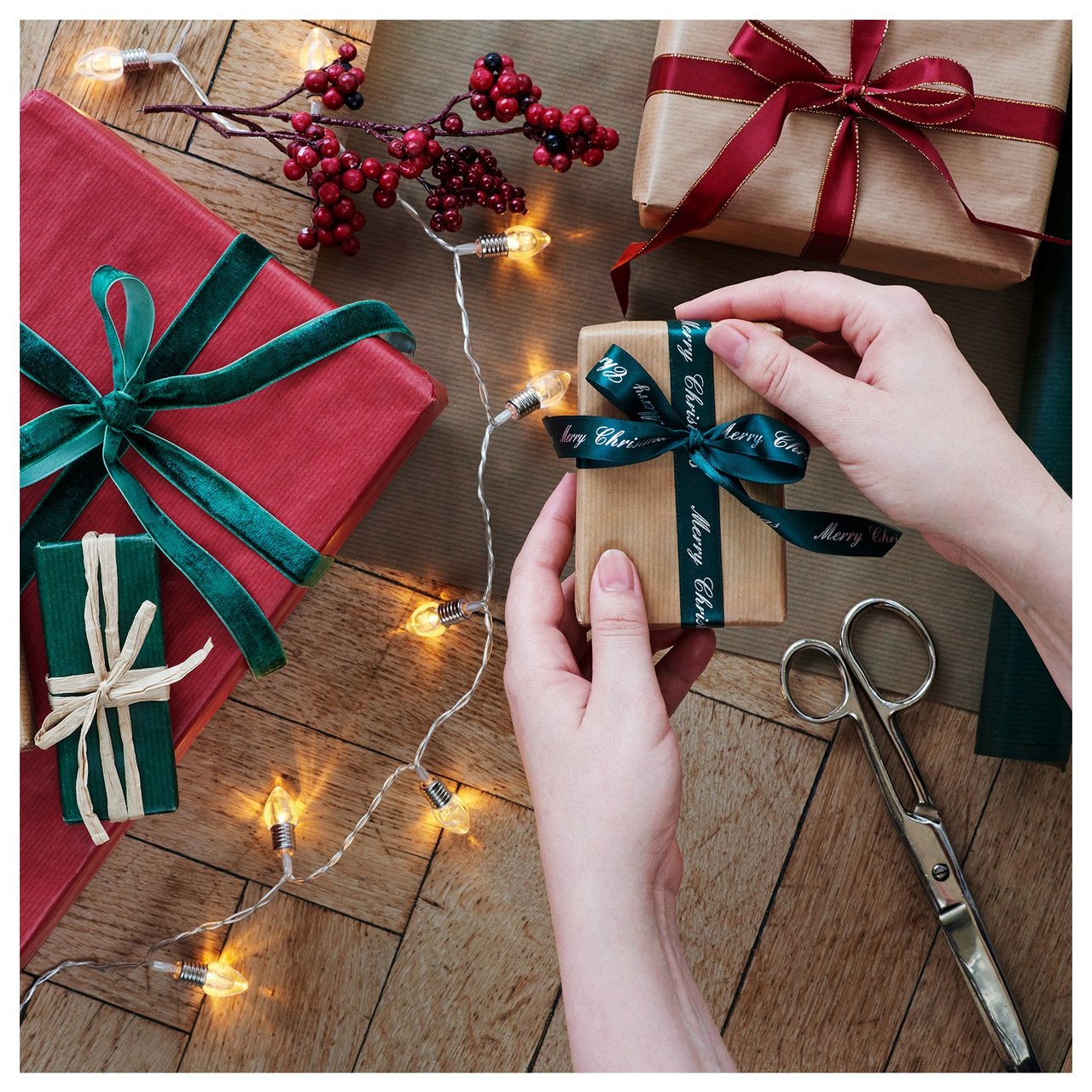 Kädet solmivat rusetin joululahjaan, ympärillä paketteja, sakset ja valoketju.