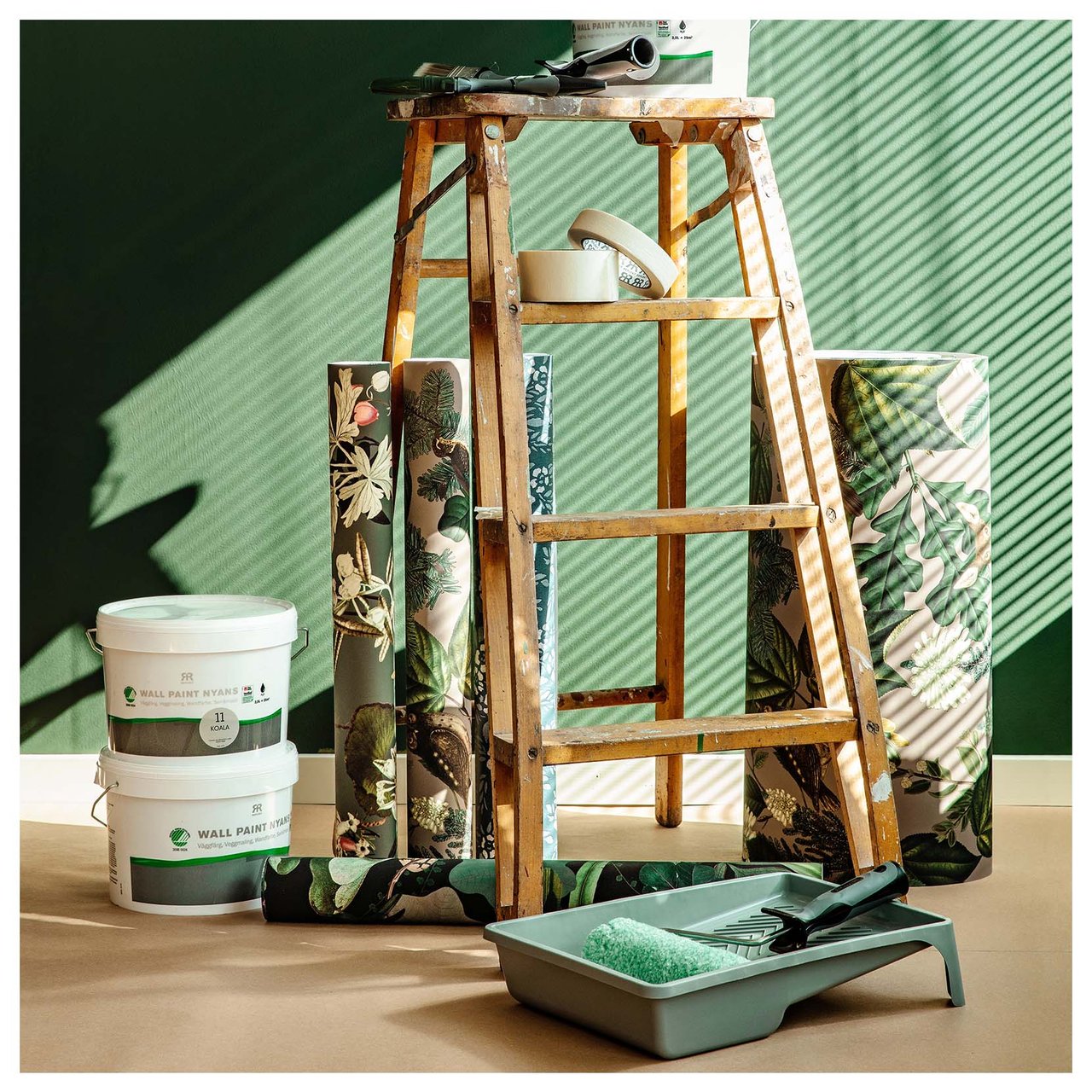 Tapettirullat, maalipurkit ja maalaustyökalut puisten tikkaiden ympärillä vihreän seinän vieressä.