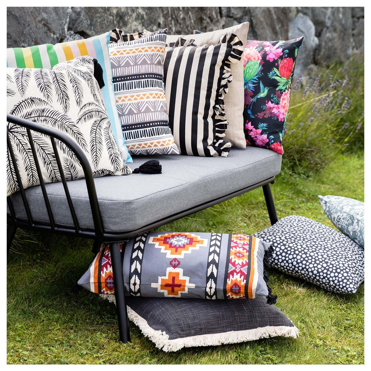 Gartensofa auf einem Rasen im Garten mit vielen Kissen in verschiedenen Farben und Mustern auf und neben dem Sofa.