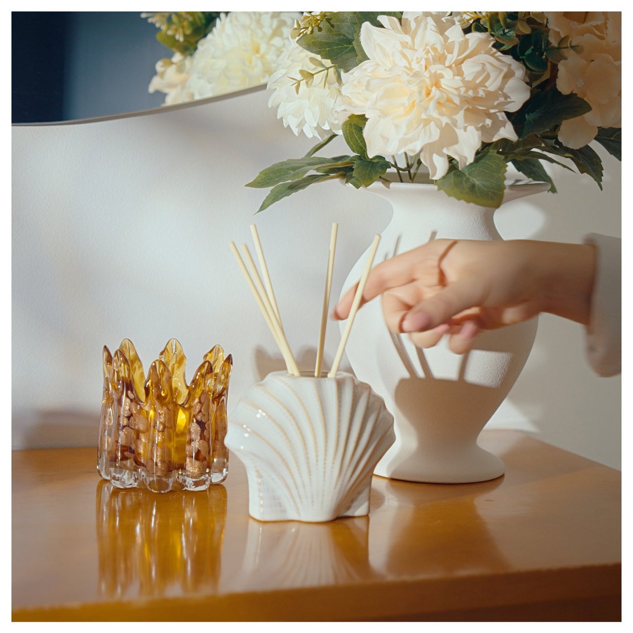 Käsi asettelee tuoksutikkuja pöydälle, jolla on kukkamaljakko ja kynttilälyhty koristeellisessa sisätilassa.