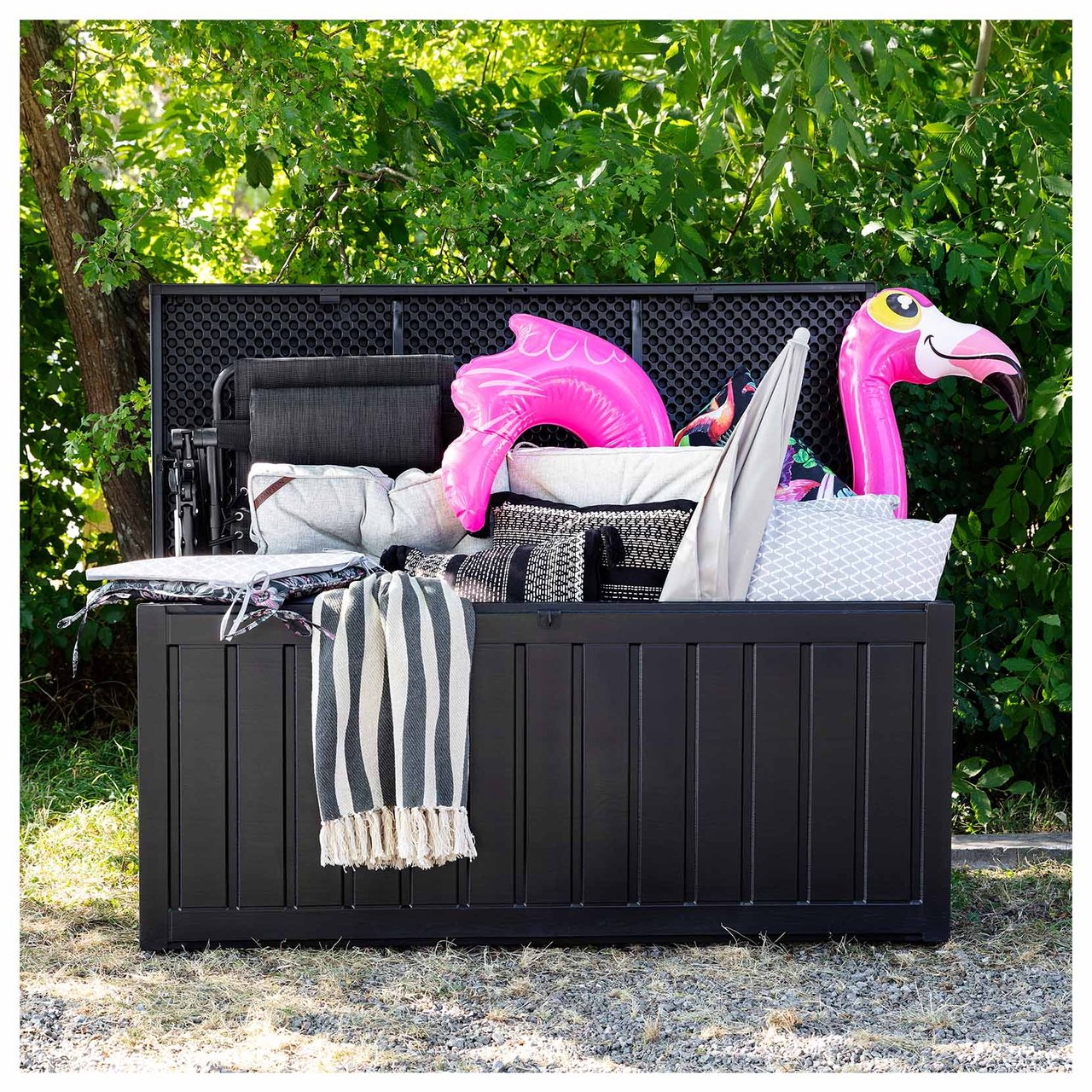 Große schwarze Auflagenbox aus Kunststoff mit offenem Deckel auf einem Rasen. In der Box befinden sich Kissen, Badespielzeug und ein Sonnenschirm.