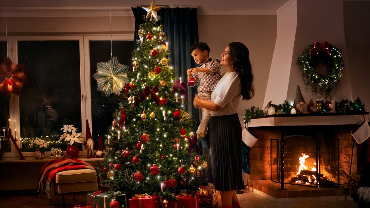 Kvinna med ett barn i famnen, bredvid en pyntad julgran med julklappar under.