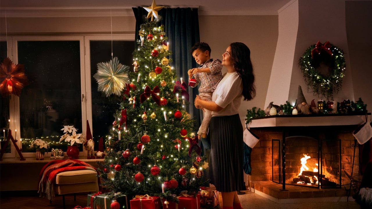 Kvinna med ett barn i famnen, bredvid en pyntad julgran med julklappar under.