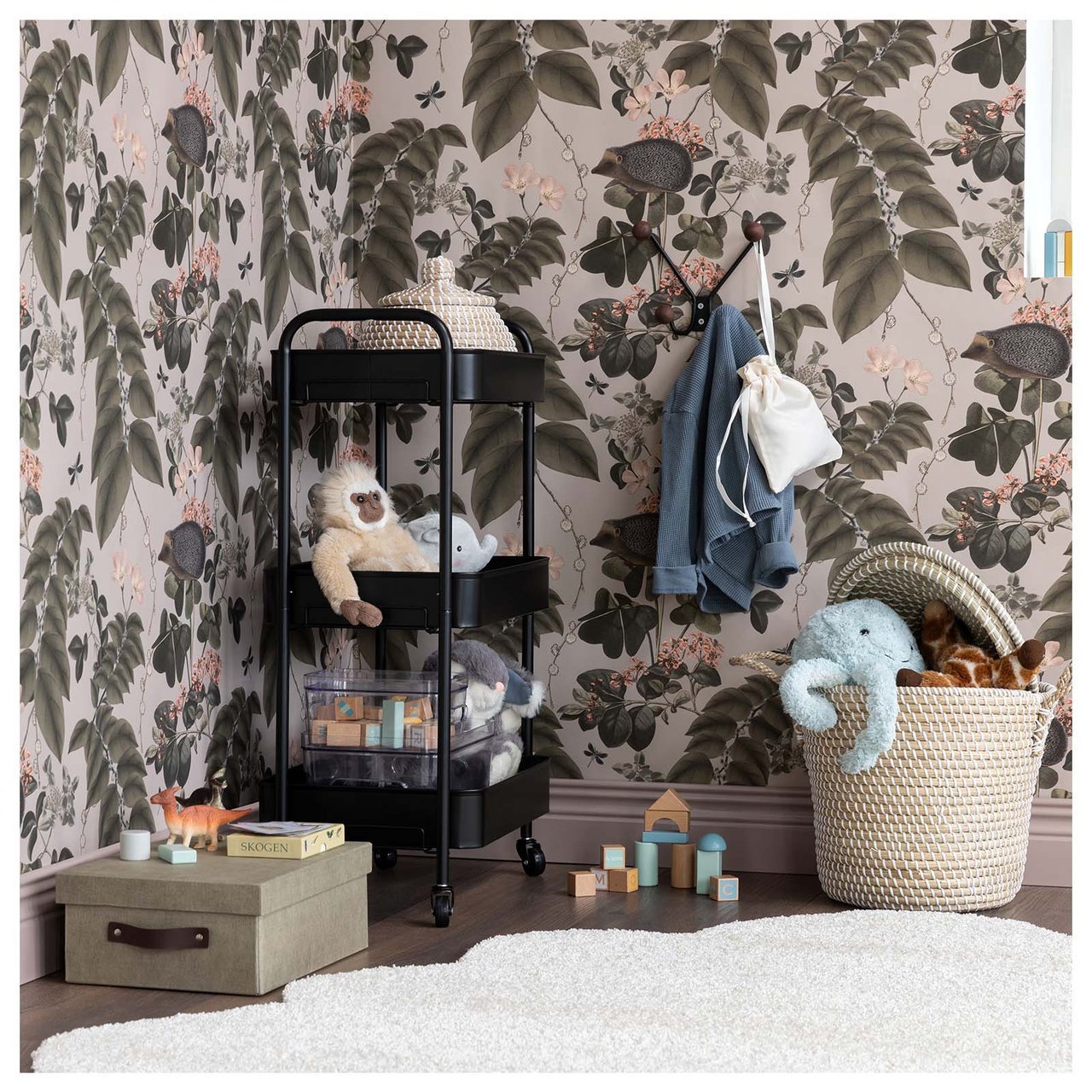 Spielzeug auf einem Rollwagen und in Körben, die auf dem Boden stehen. Im Hintergrund eine dekorative Tapete mit Blattmuster.