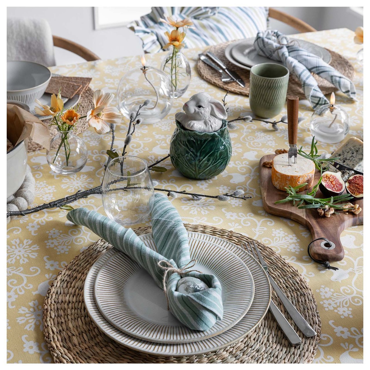 Gedeckter Ostertisch mit Platzdeckchen, Geschirr, Stoffservietten, Käseplatte und einer Duftkerze in Hasenform.