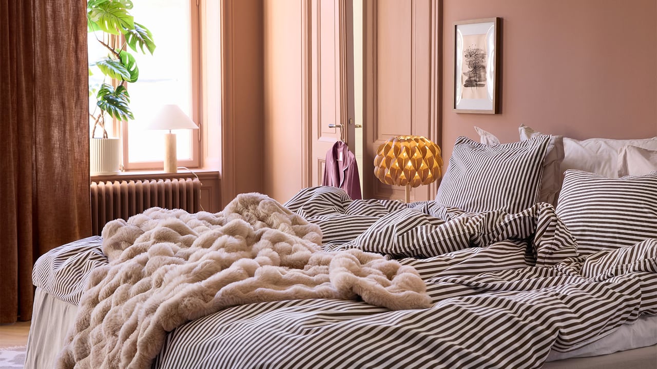 Ett sovrum med terrakottafärgade väggar, randiga sängkläder och en fluffig, beige pläd.