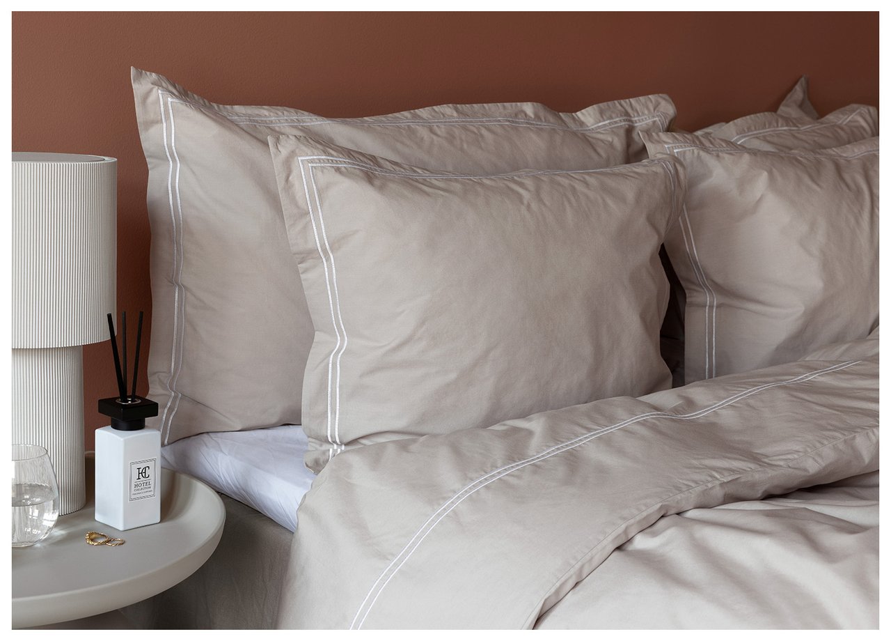 Hodeenden av en seng med store hotellputer bakerst og vanlige puter foran. Beige sengetøy med dekorativ hvit rand.