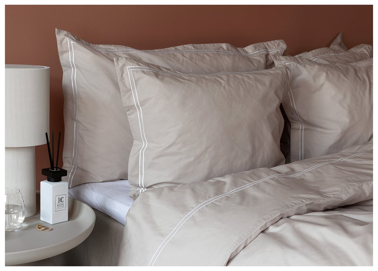 Hodeenden av en seng med store hotellputer bakerst og vanlige puter foran. Beige sengetøy med dekorativ hvit rand.