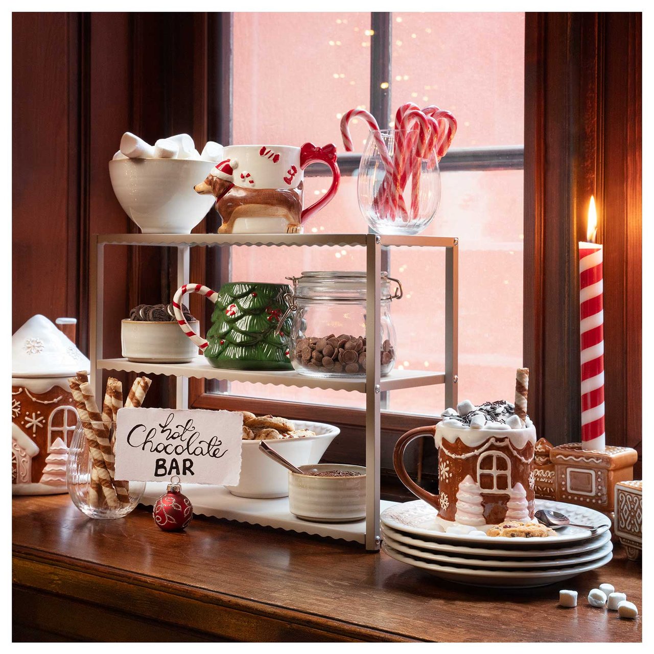 En hot chocolate bar uppdukad i ett fönster med julmuggar, marshmallows, polkagrisar och tända ljus.