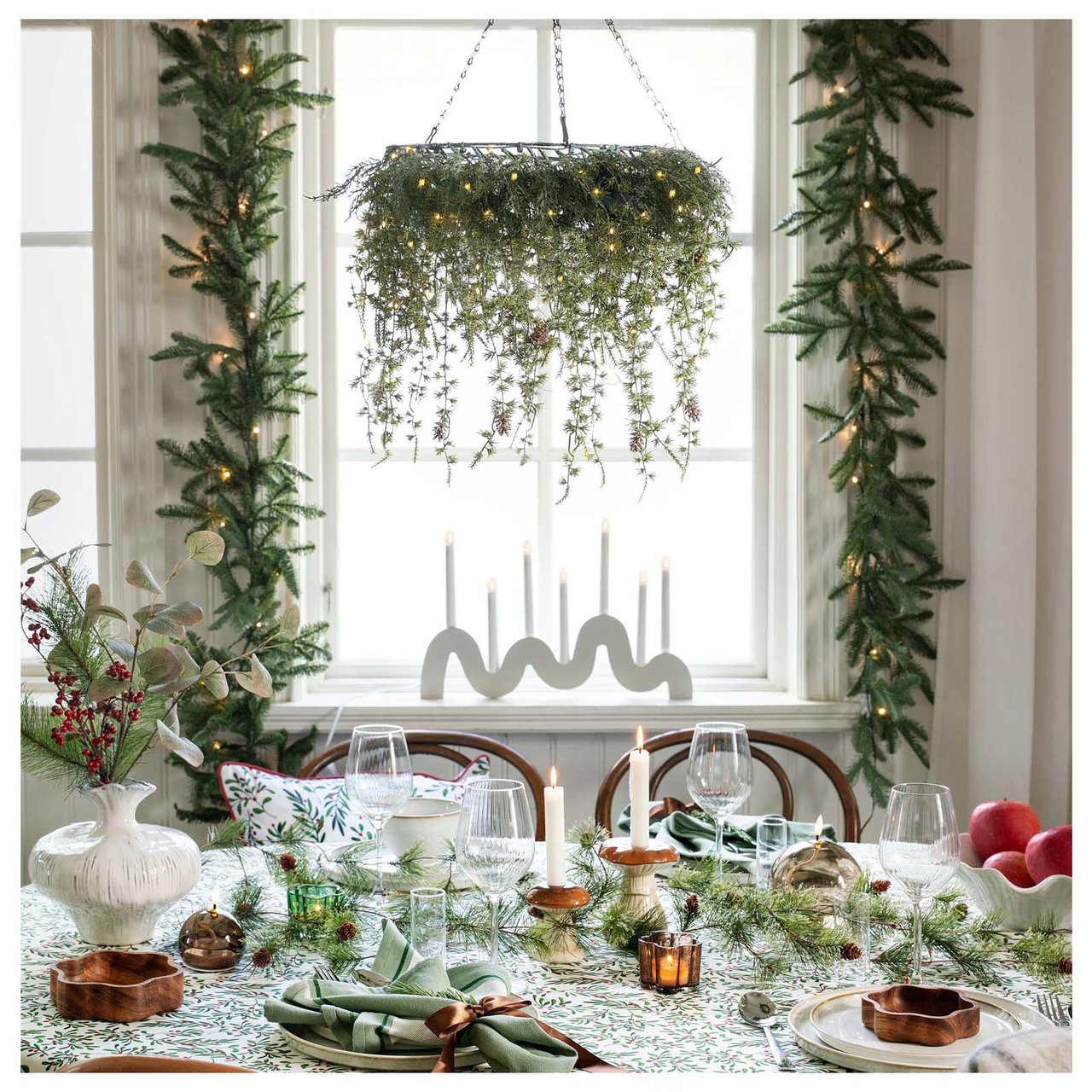 Ein Esstisch, in Grün und Weiß gedeckt, steht vor einem mit Tannenzweigen geschmückten Fenster.
