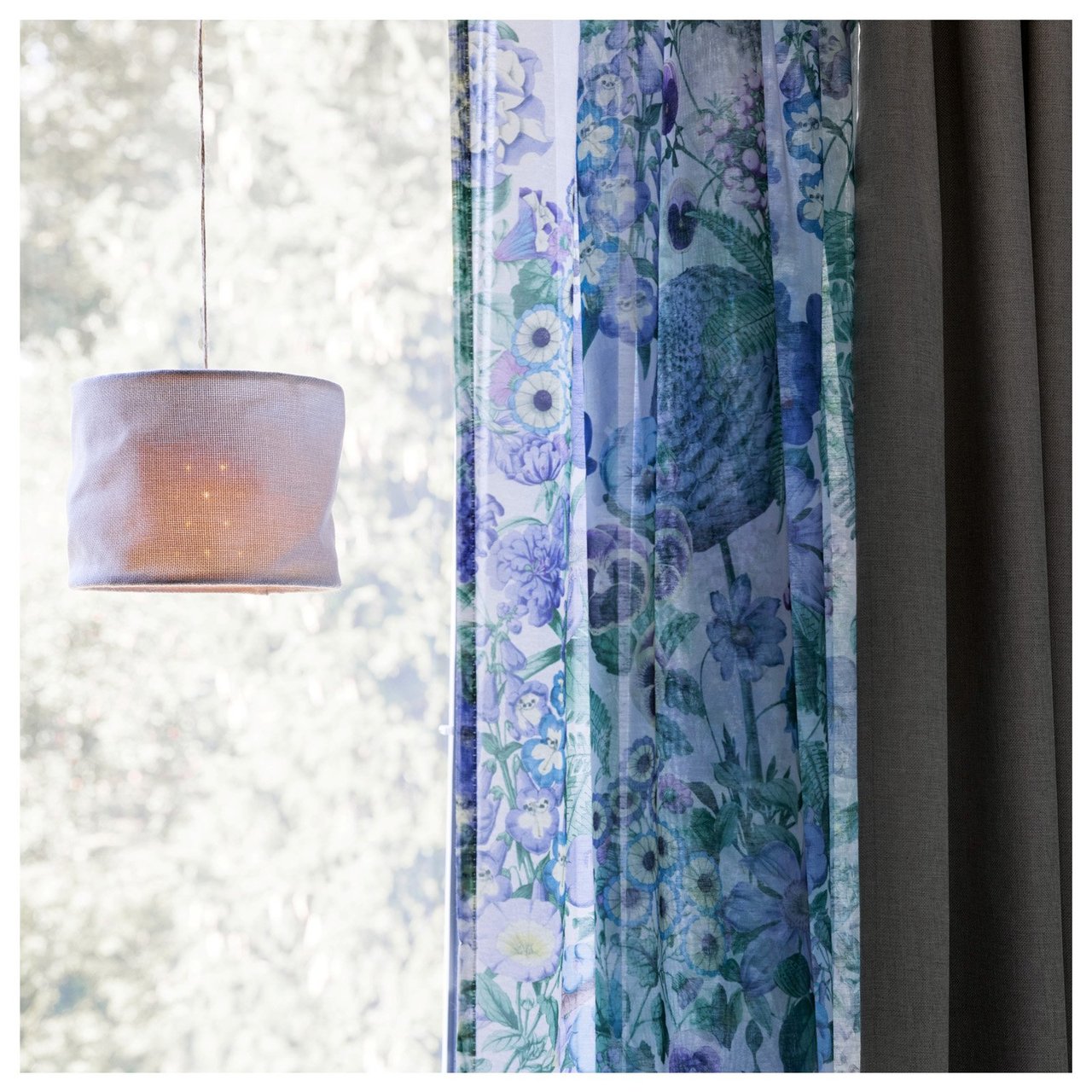 Deckenlampe aus Textil, die vor einem sonnendurchfluteten Fenster mit geblümten und einfarbigen Vorhängen hängt.