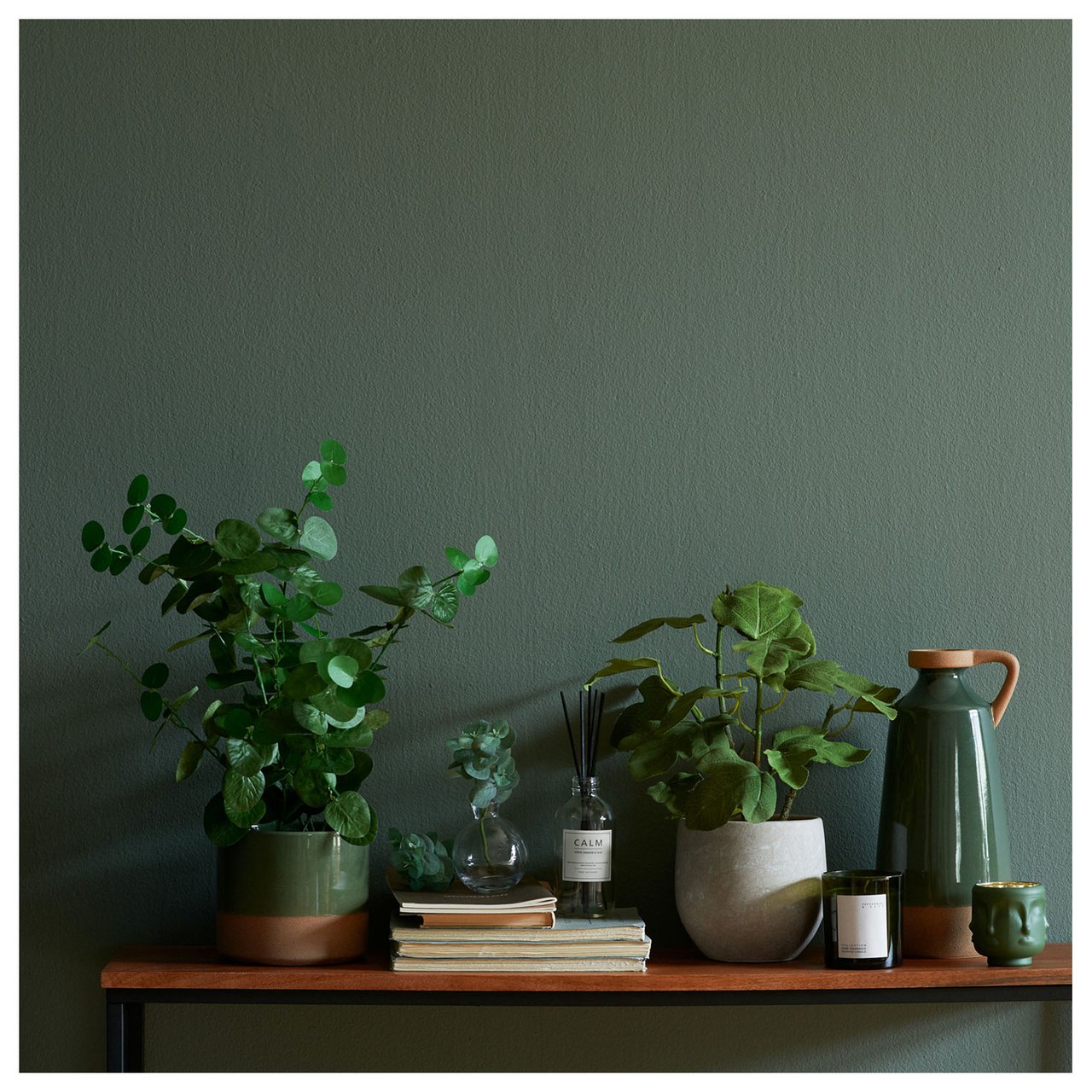 Hylle med grønne planter, duftpinner, bøker og dekorative detaljer mot en grønn vegg.