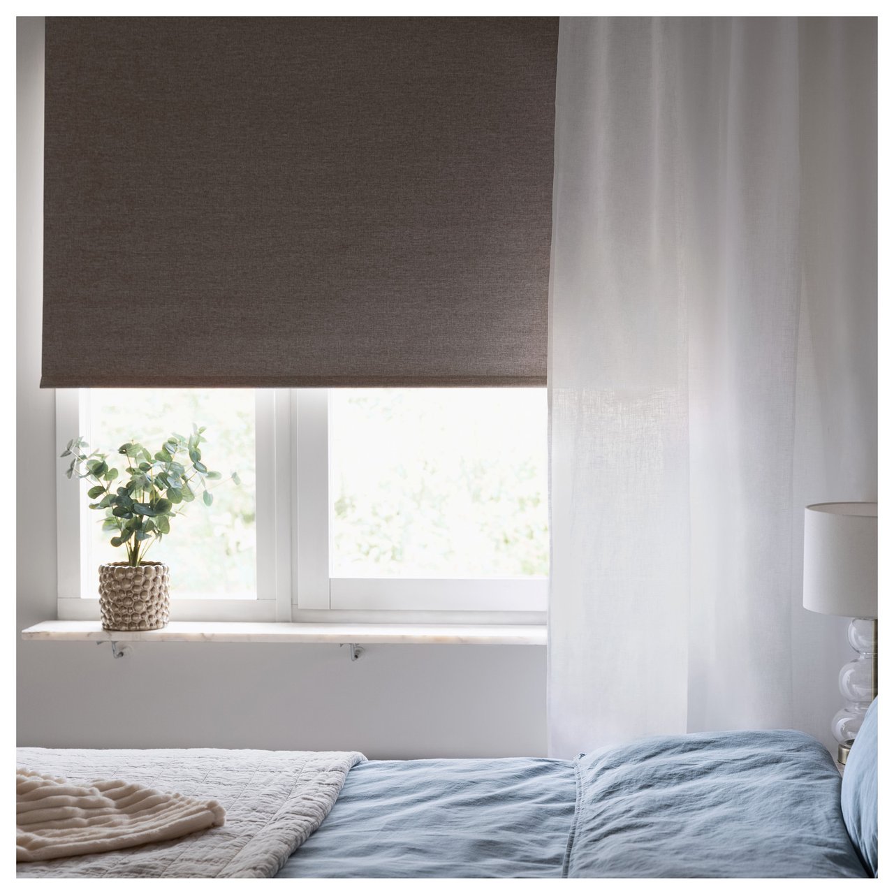 Bett am Fenster mit Rollo und durchsichtigen Vorhängen, Tischleuchte und Pflanze am Fenster.