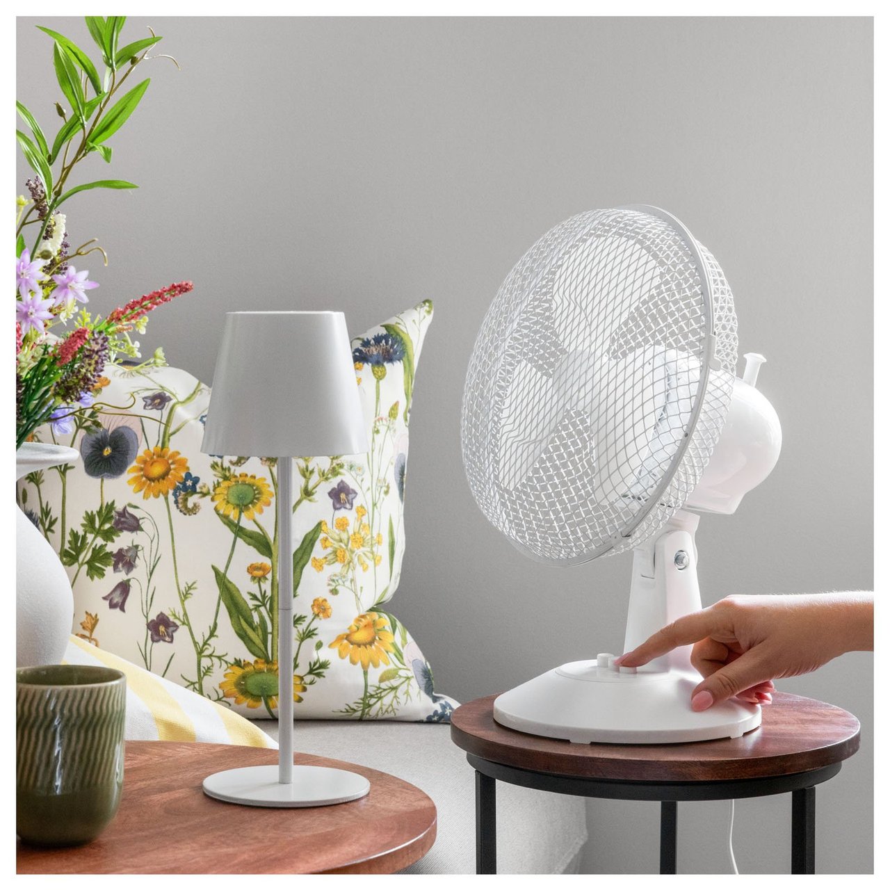 En person startar en bordsfläkt i ett rum med blomkudde, lampa och dekorativa detaljer.