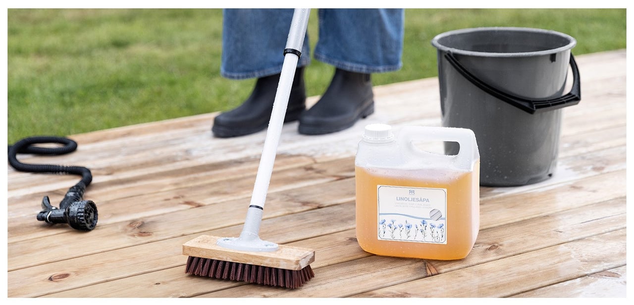 Eine Person reinigt eine Holzterrasse mit Leinölseife und einer Bürste. Auch ein Eimer und ein Gartenschlauch sind zu sehen.