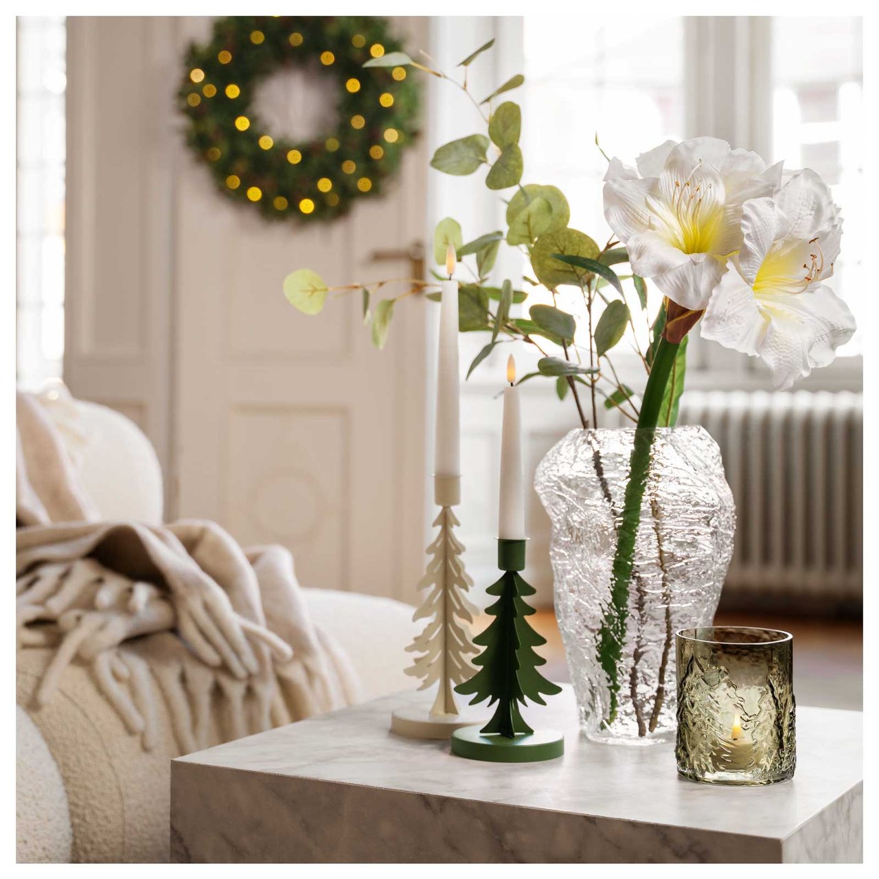 Glasvase mit weißen Amaryllis und Tannenbaum-Kerzenhaltern auf einem Couchtisch. Im Hintergrund eine Tür mit leuchtendem Kranz.