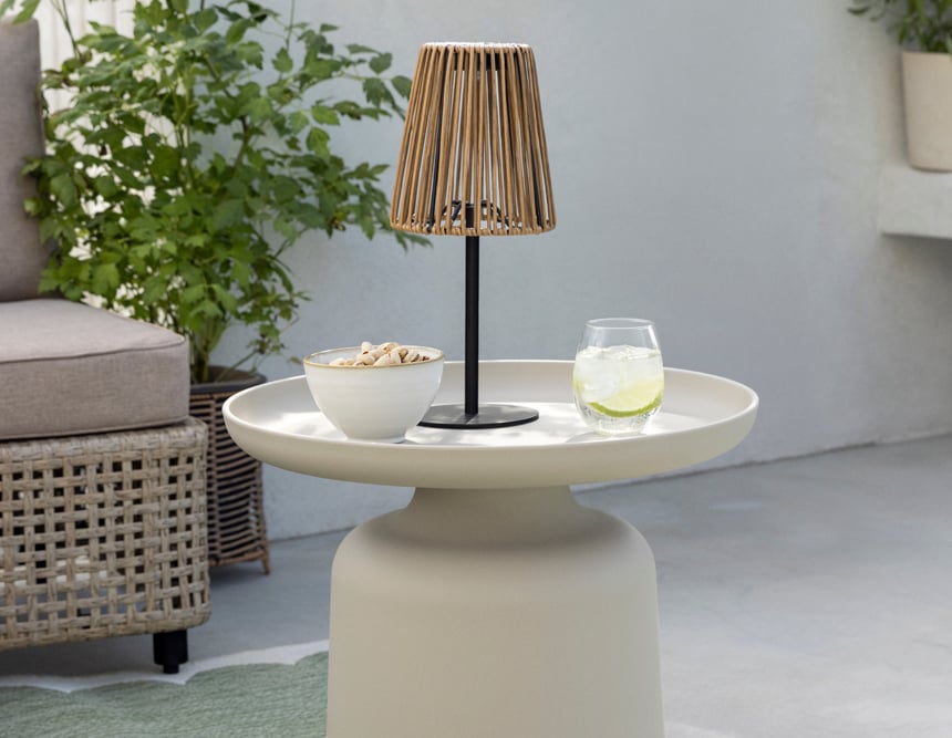 Runder Tisch mit Lampe, Snacks und Getränk auf Terrasse mit Pflanzen und Loungemöbeln.