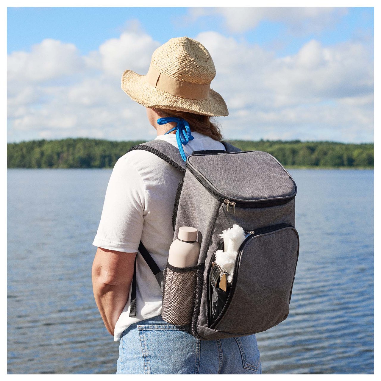 Eine Person mit Strohhut und praktischem Kühlrucksack blickt über einen See, bereit für einen schönen Ausflug mit einer Wasserflasche und Picknickaccessoires.