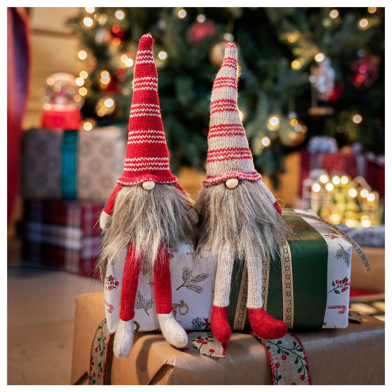Zwei kleine Weihnachtsmänner sitzen auf Weihnachtsgeschenken vor einem geschmückten Weihnachtsbaum.