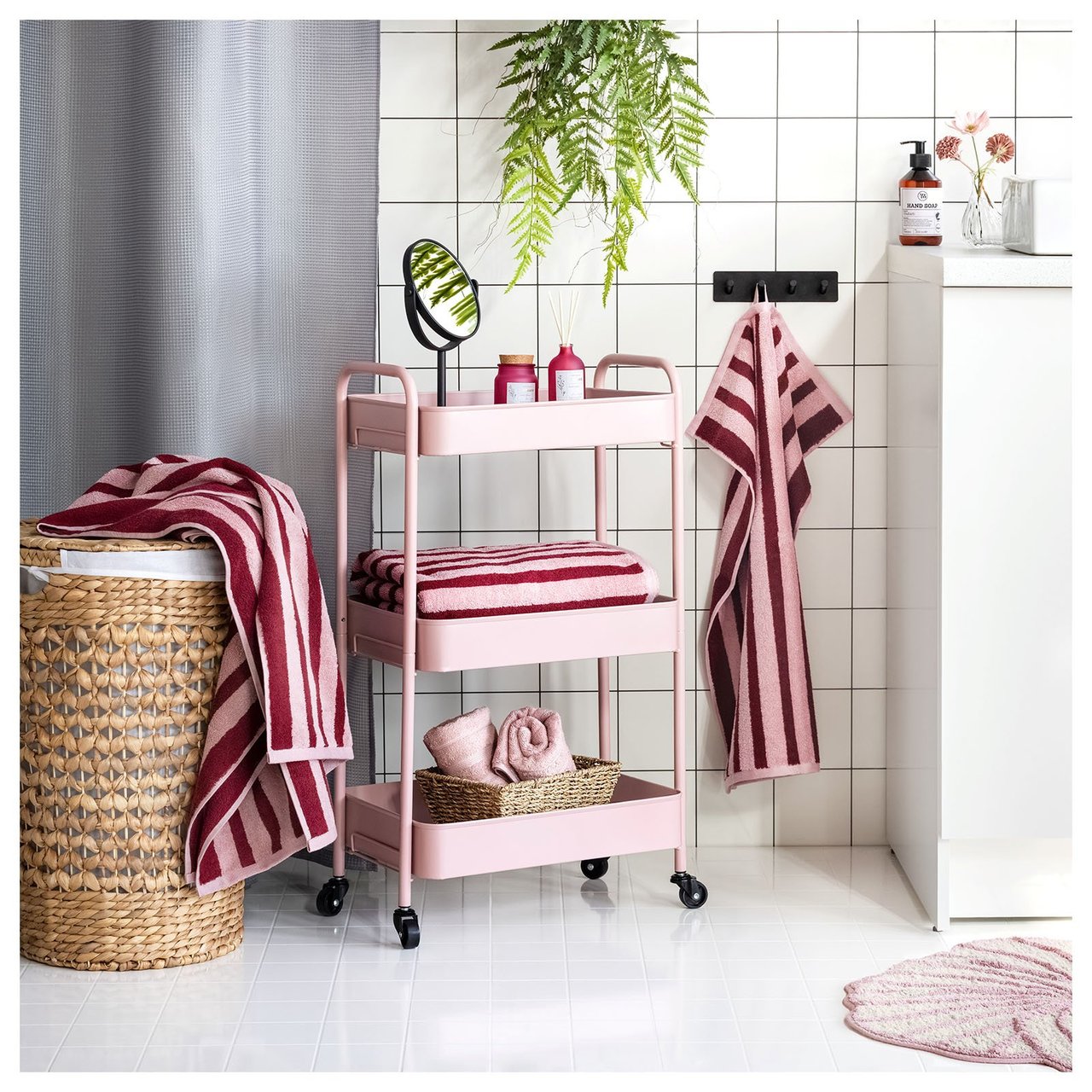 Weiß gefliestes Badezimmer mit Rollwagen, Handtüchern und Badematte in Rosa. Wäschekorb aus Rattan und schwarzer Toilettenpapierhalter.