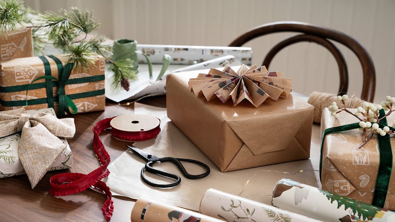 Geschenke werden mit braunem Papier, rotem Band und einer Tannenzweig auf dem Tisch verpackt.